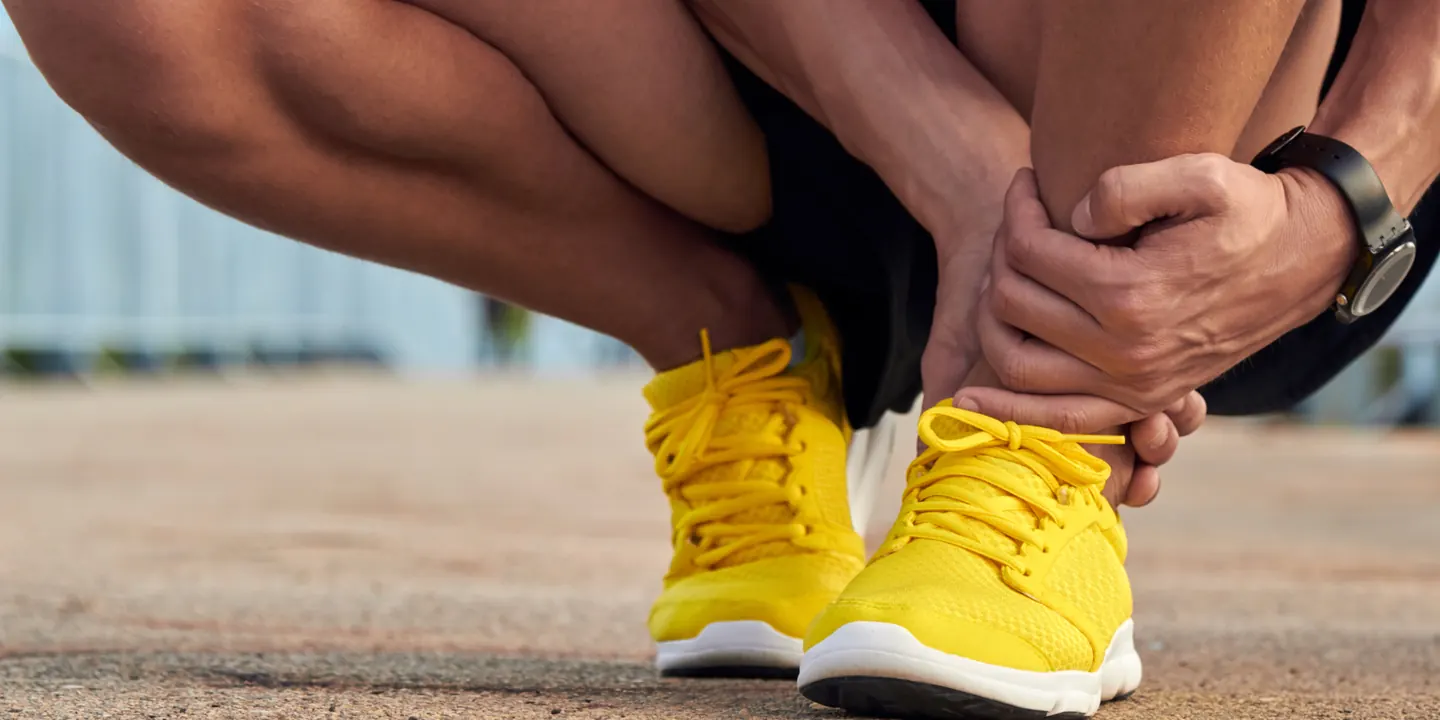 Runner in yellow trainers crouching on a path, gripping the back of the ankle where the Achilles tendon hurts